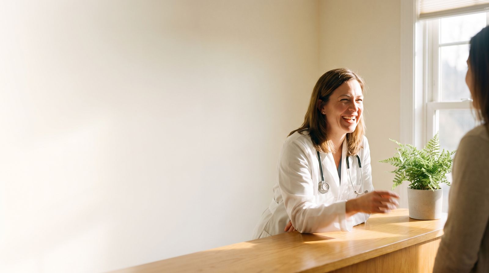 A physician in a white coat laughing mid-conversation at a sunlit counter with a fern beside her.